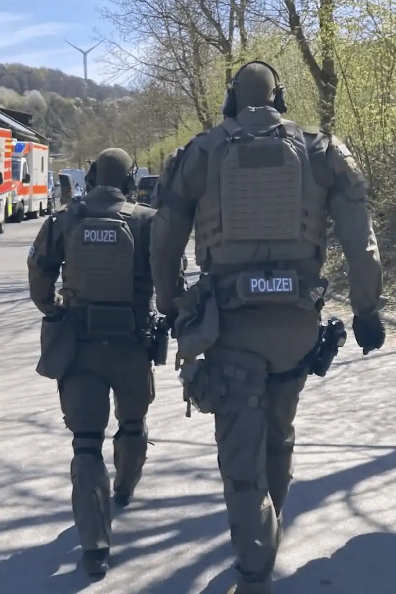 Two German police officers in tactical gear walk away from the camera down a paved road, their vests clearly labeled…
