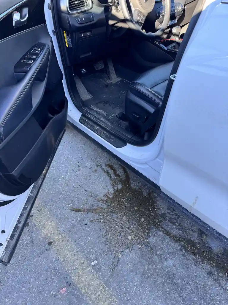 A white car's driver-side door is open, revealing a dirty interior with a black floor mat. On the concrete ground outside…