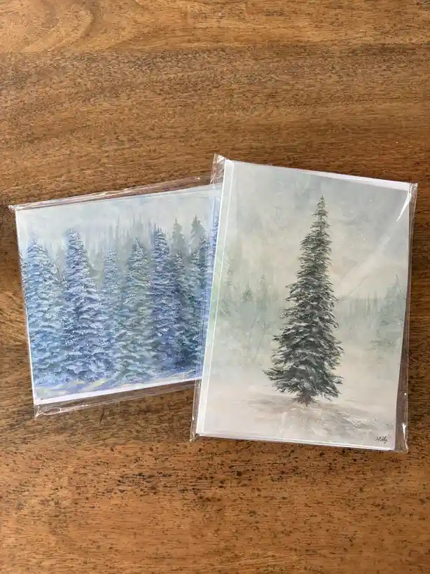 Two Christmas cards in clear plastic sleeves lie on a wooden surface. One card depicts a dense forest of snow-covered pine…