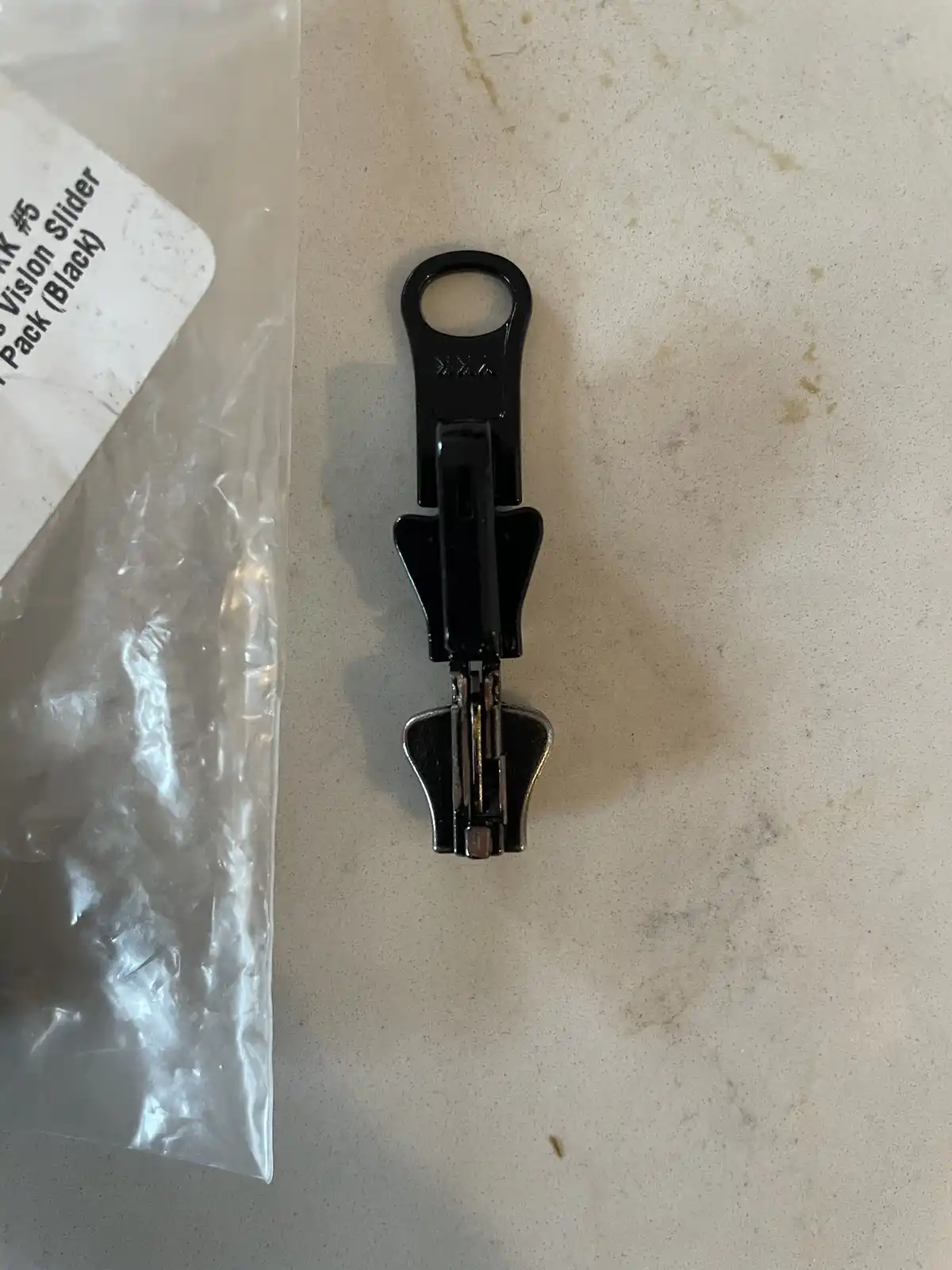 A black zipper slider lies on a speckled countertop next to its clear plastic packaging, which has visible text including…
