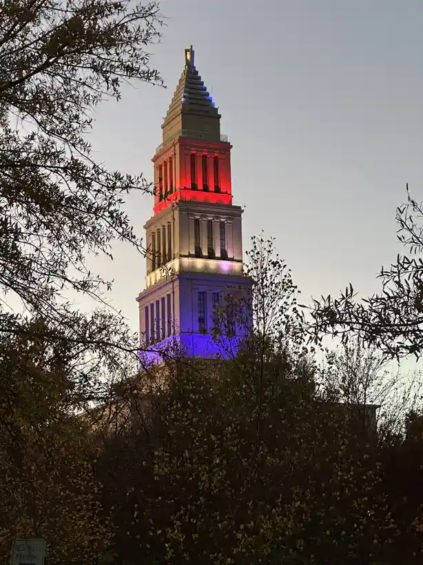 A tall, multi-tiered stone tower is illuminated at dusk with red lights on the upper section, white in the middle, and blue…