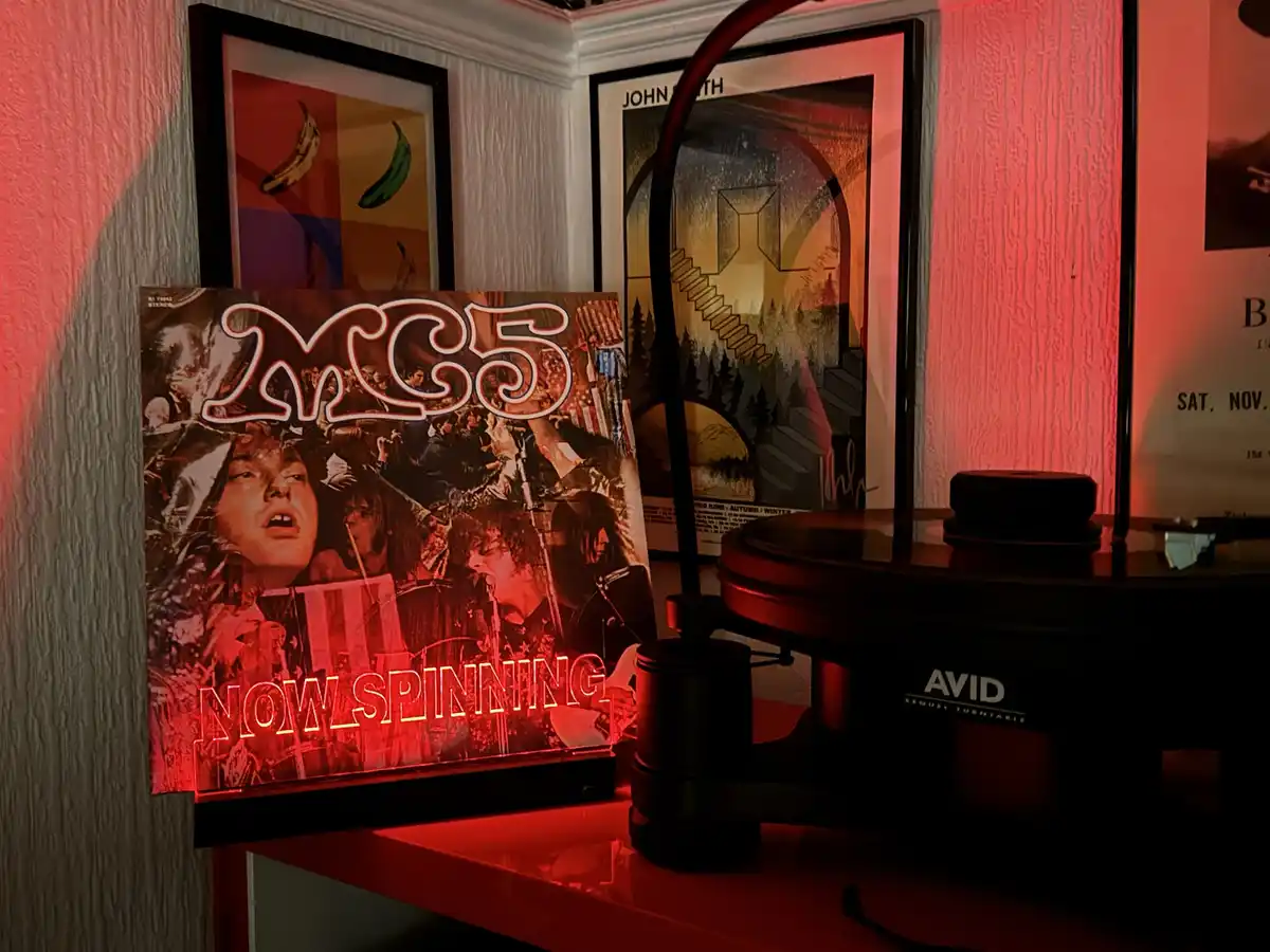 A red-lit room features a vinyl record player with an 'AVID' brand turntable. Next to it, a glowing neon sign displays the…