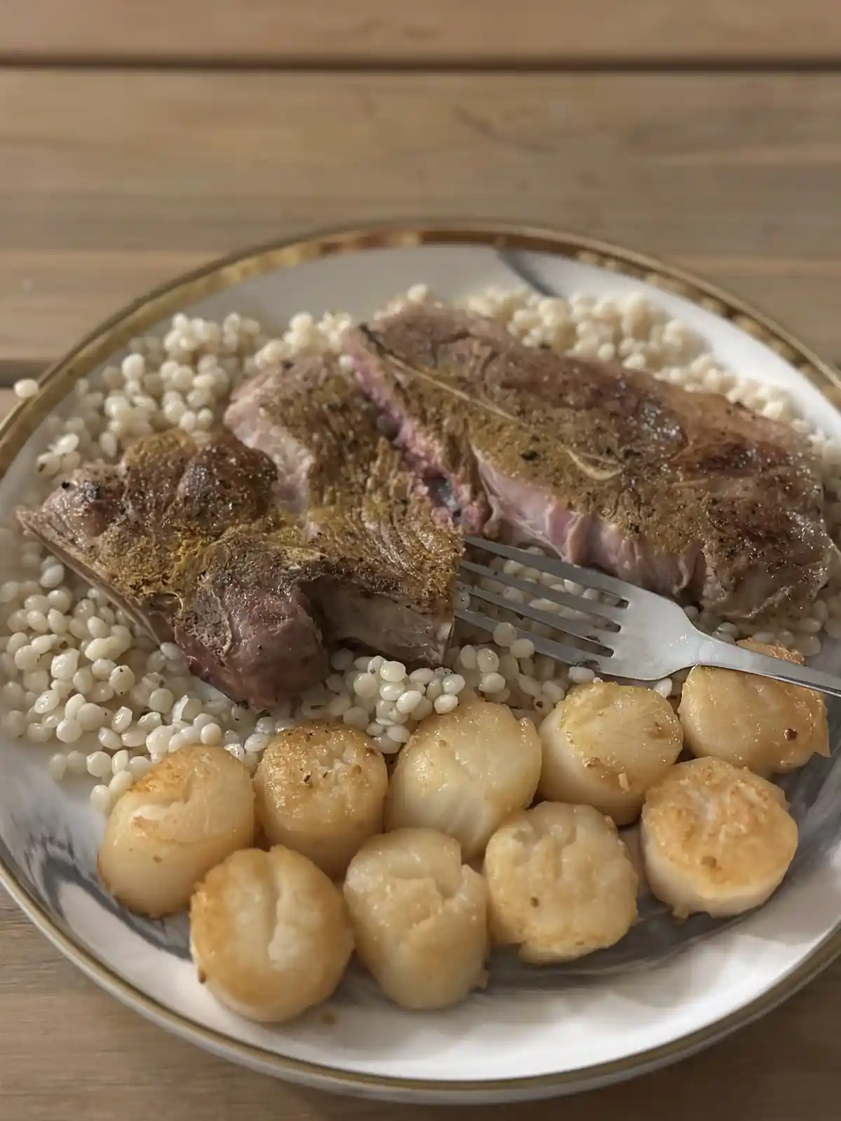 A close-up photo of a plated meal featuring a seared meat cut (possibly lamb or pork chop) resting on a bed of small white…
