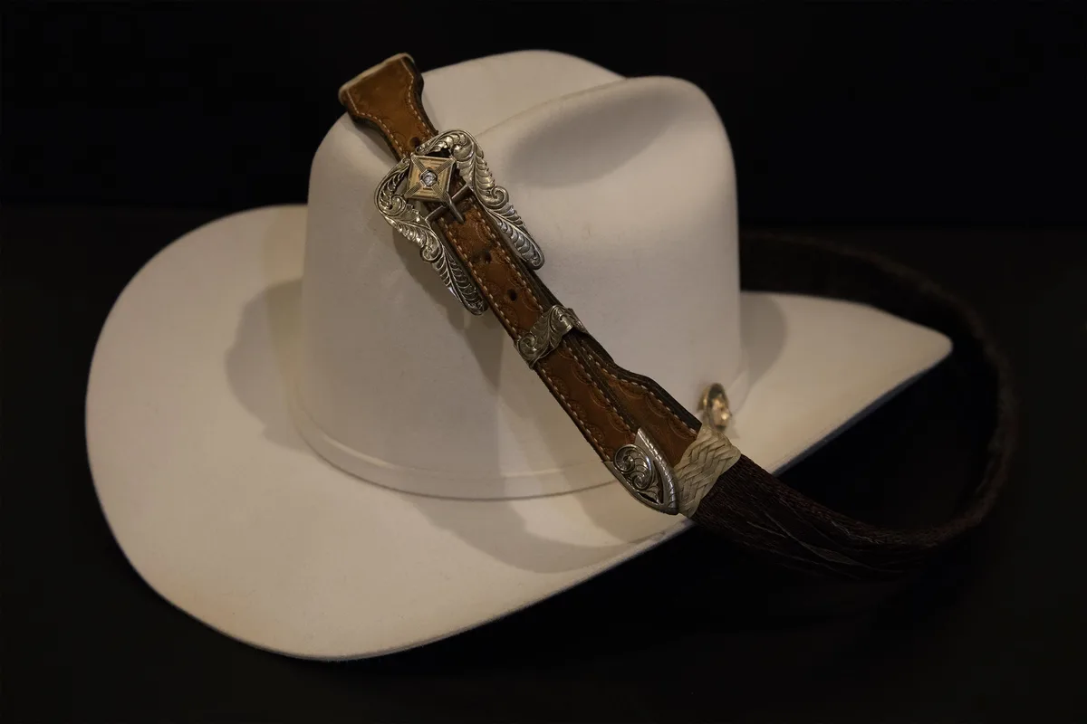 A white cowboy hat with a decorative brown leather strap and ornate silver conchos is displayed against a black background…