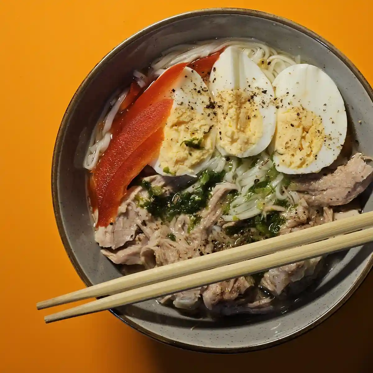 A top-down photo of a bowl of ramen with white noodles, sliced boiled eggs, shredded meat, green herbs, and a slice of red…