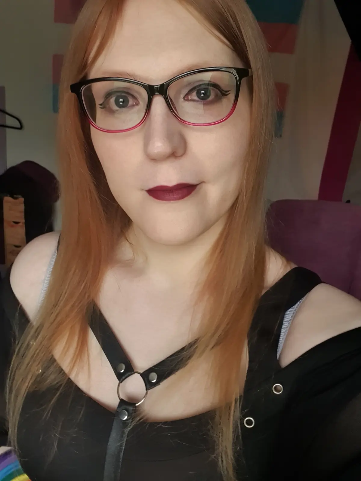 A person with long reddish-brown hair and black-rimmed glasses with pink inner edges stares directly at the camera with a…
