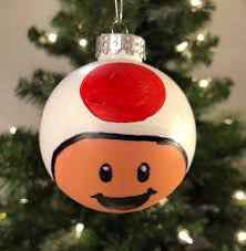 A hand-painted Christmas ornament shaped like Toad from Mario, featuring his iconic white mushroom cap with a red spot and…