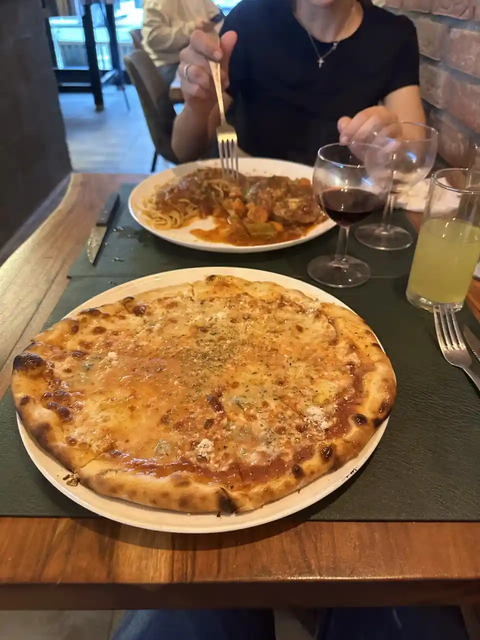 A person sits at a restaurant table with a large cheese pizza in front of them, while another plate with pasta and meat is…