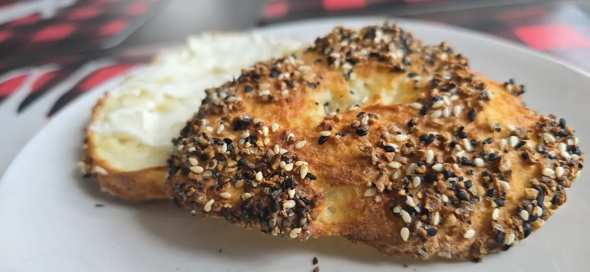 A close-up photo shows a sliced bagel topped with black and white sesame seeds and other grains, with a generous spread of…