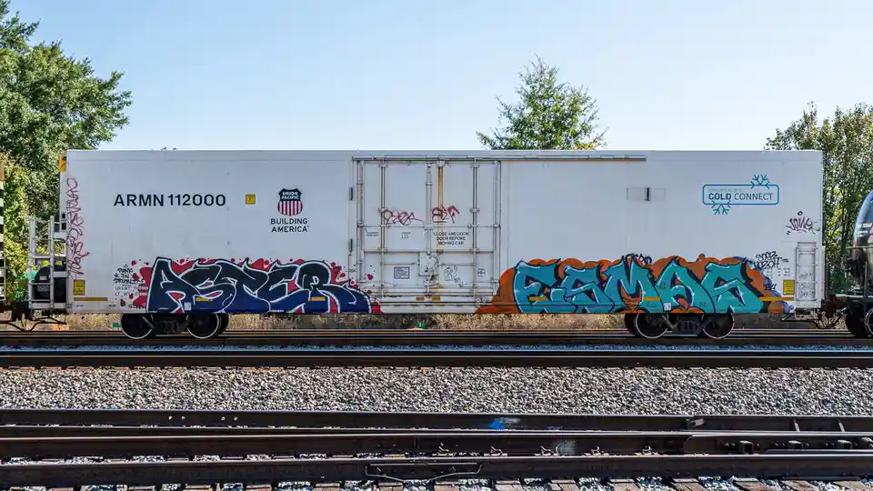 A white Union Pacific refrigerated railcar, marked with 'ARMN 112000' and 'Building America', sits on tracks under a clear…