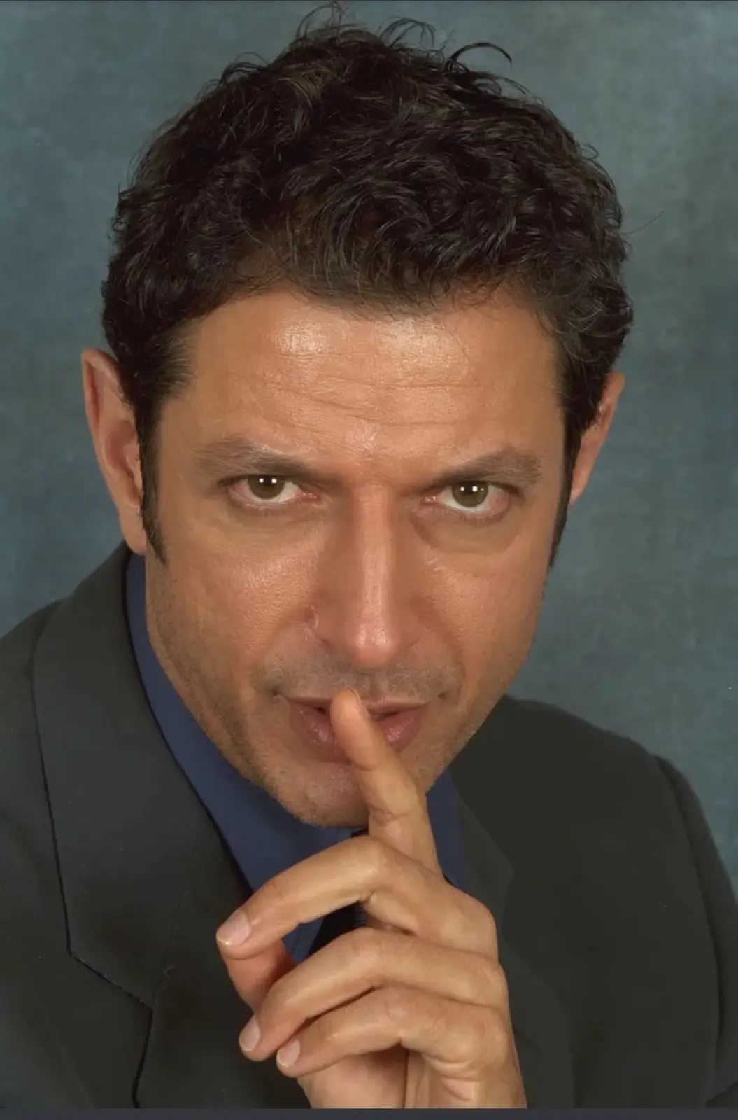 A man with dark curly hair and a serious expression, wearing a dark suit and blue shirt, holds his index finger vertically…