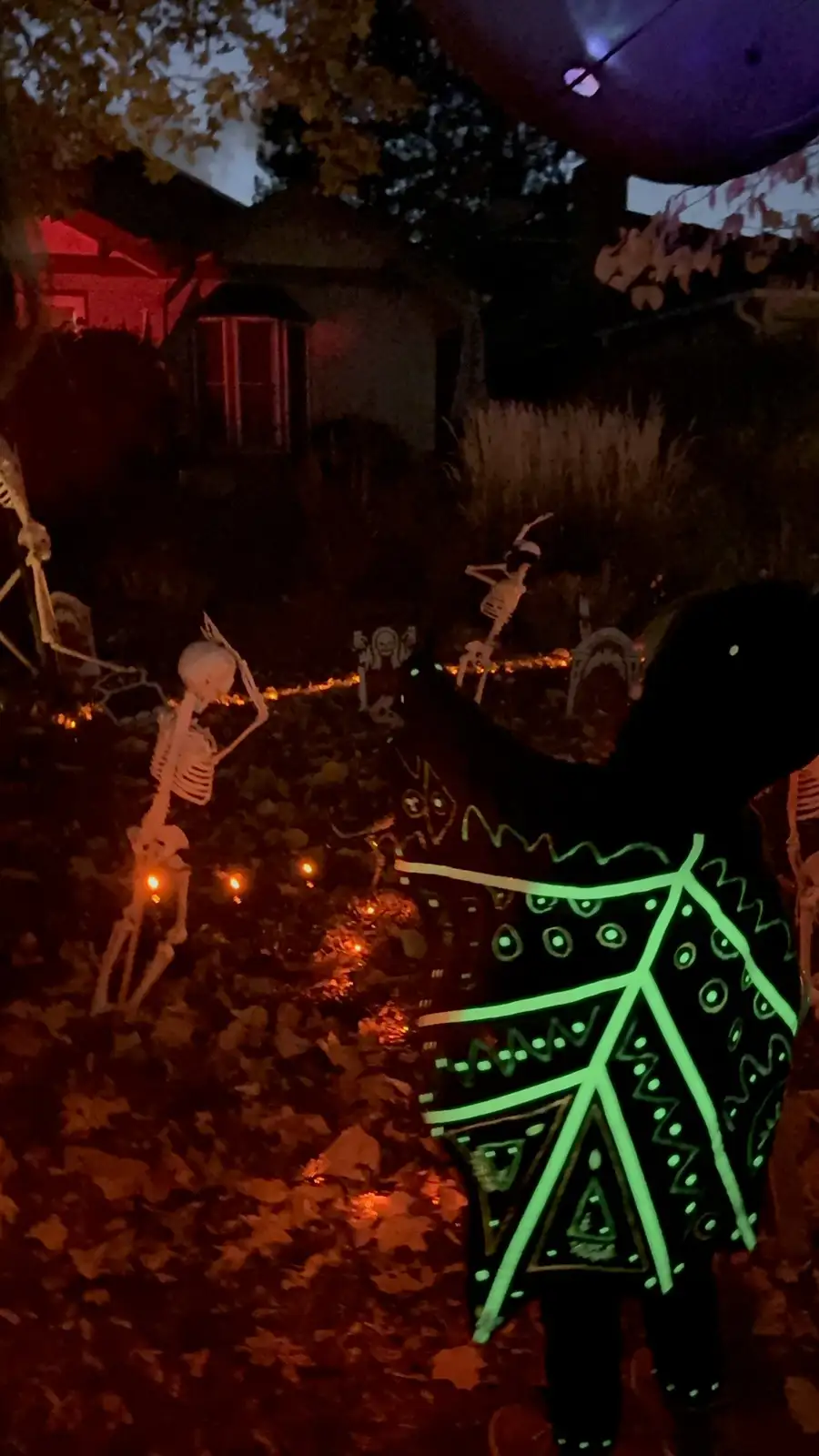 A person wearing a black outfit with bright green glowing tribal patterns stands in a dark yard at night, surrounded by…