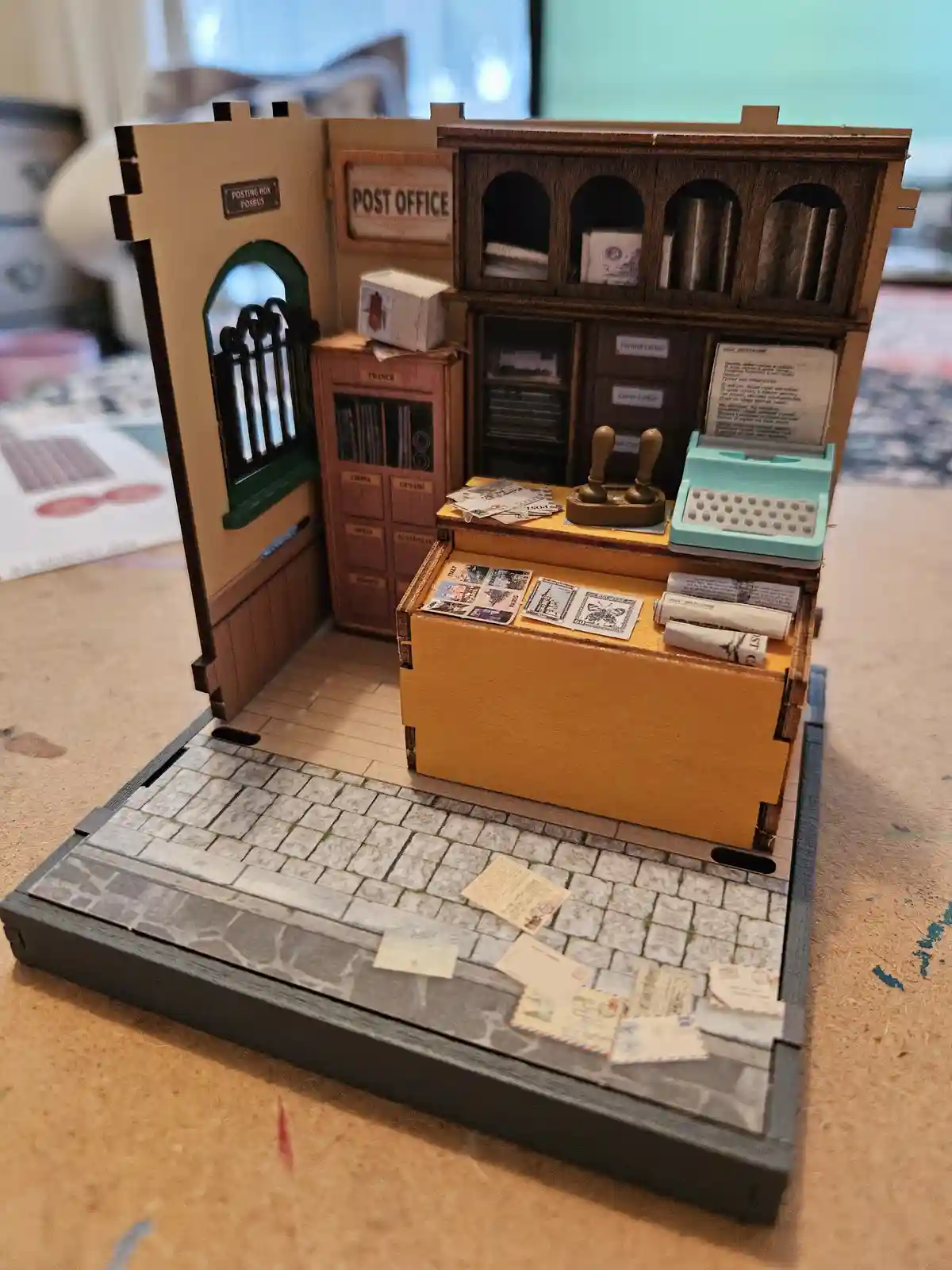 A detailed miniature diorama of a vintage post office with wooden counters, mailboxes, stamps, and a typewriter, set on a…