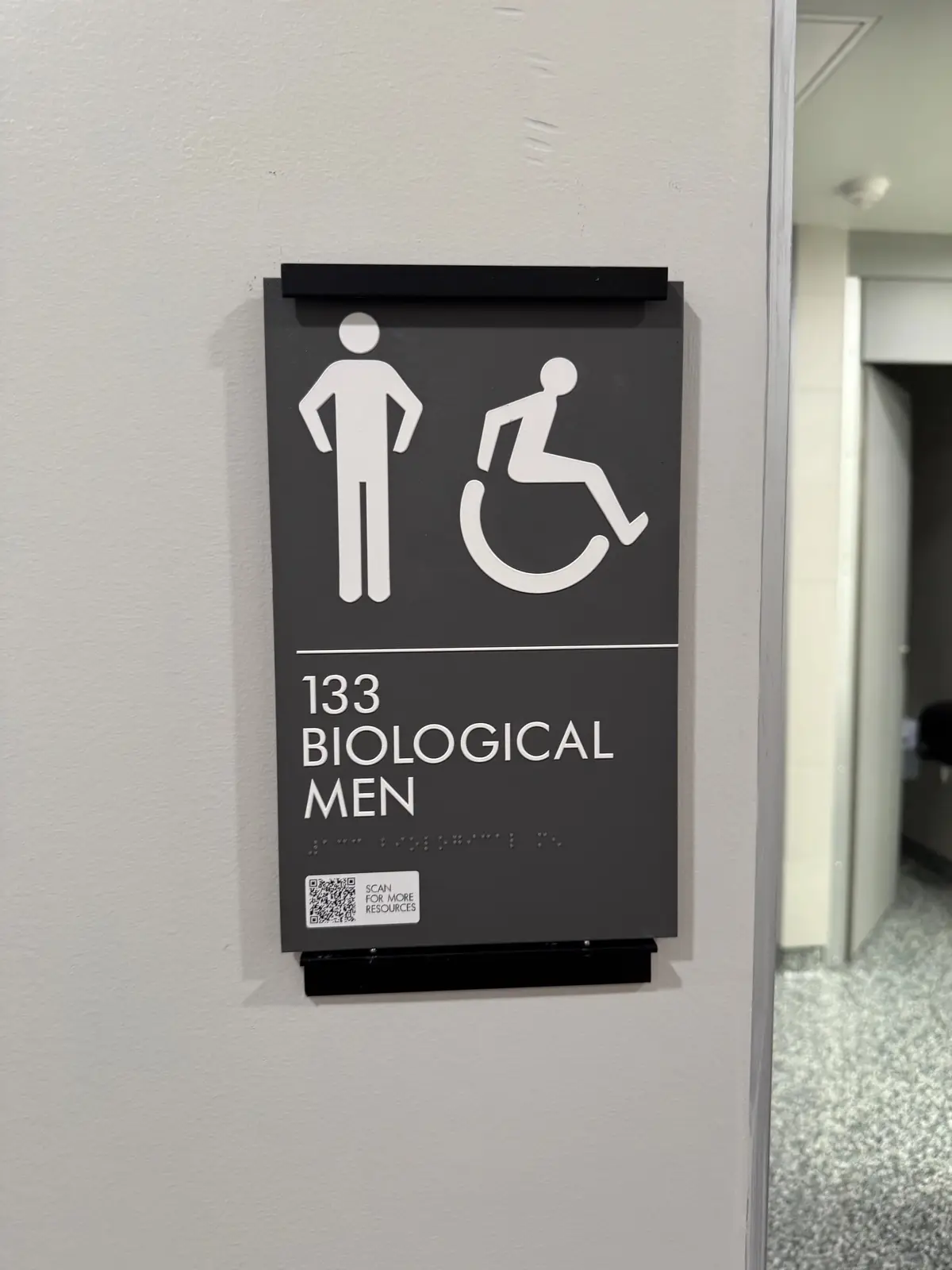 A dark gray restroom sign mounted on a white wall shows a male figure and wheelchair symbol above the text '133 BIOLOGICAL…
