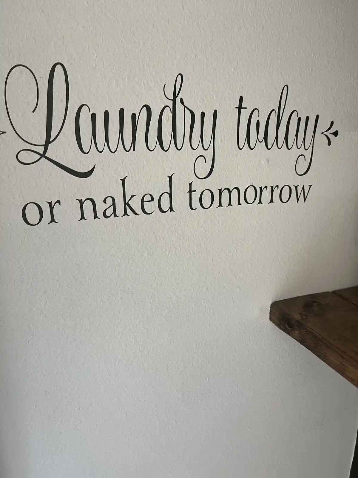 A close-up of a white textured wall with black cursive wall art that reads 'Laundry today or naked tomorrow'. The bottom…