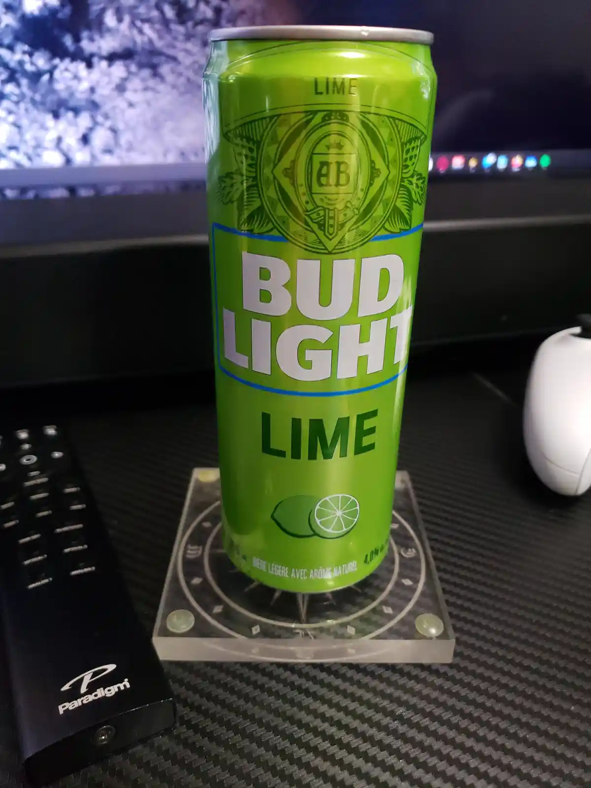 A bright green Bud Light Lime beer can sits upright on a clear acrylic coaster on a dark carbon fiber desk. To the left is a…