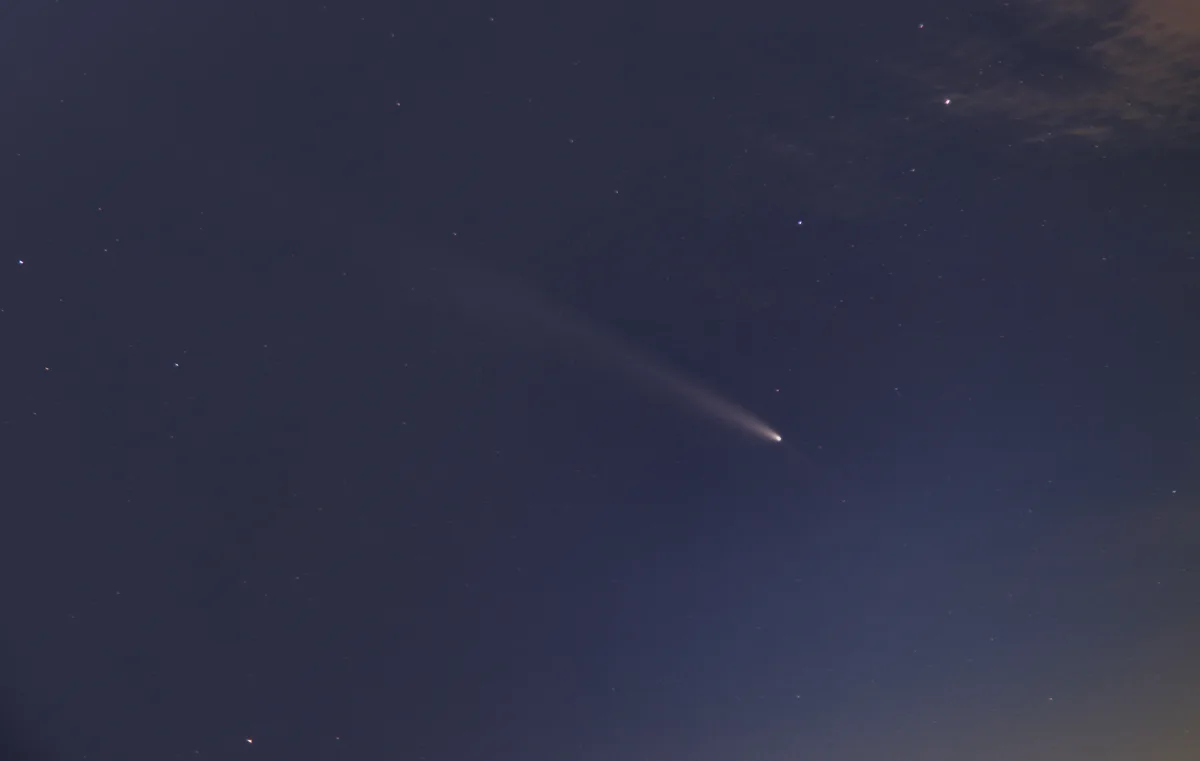 A night sky photograph shows a bright comet with a long, diffuse tail streaking diagonally across a field of stars, with…