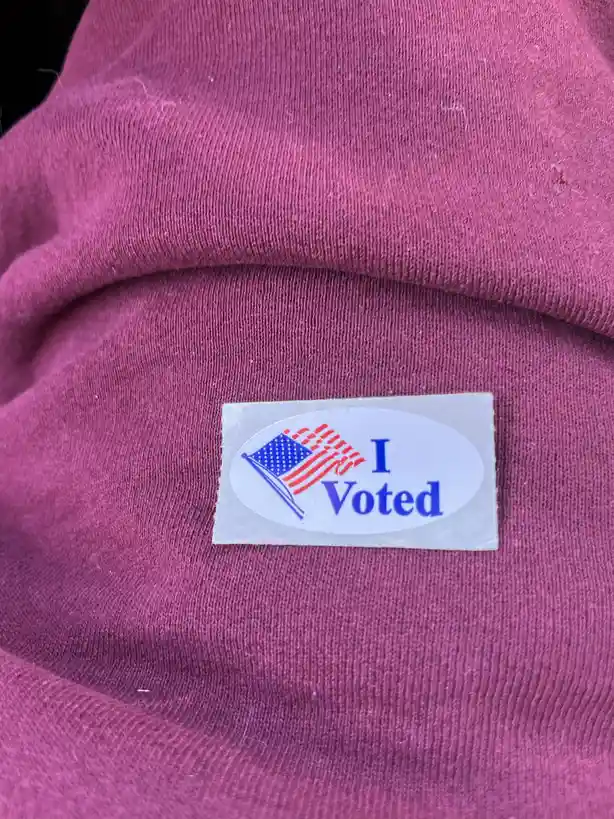 A close-up photo shows a standard American 'I Voted' sticker with a flag graphic, stuck on a maroon-colored knit garment…