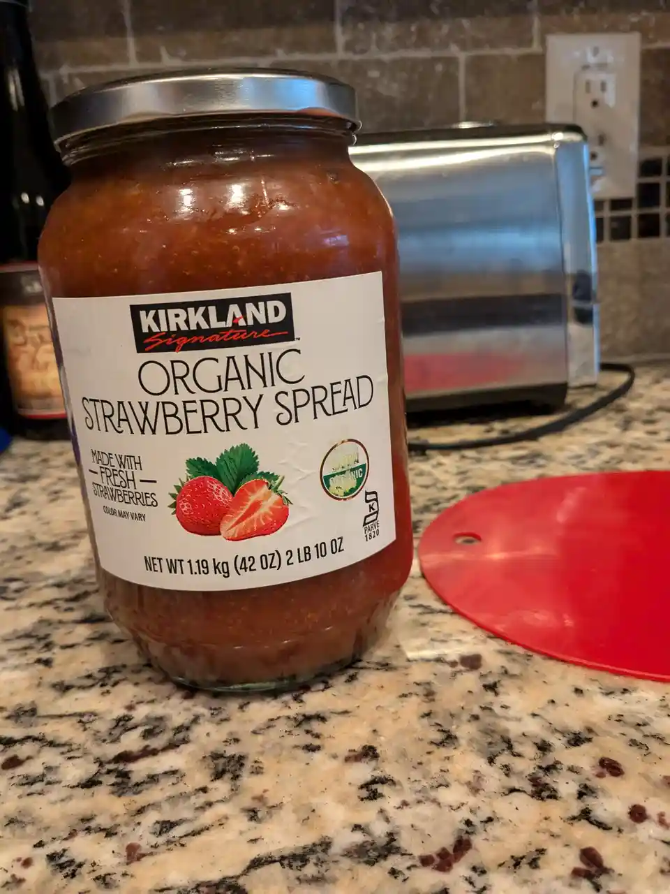 A large glass jar of Kirkland Signature Organic Strawberry Spread sits on a granite kitchen counter, with a stainless steel…