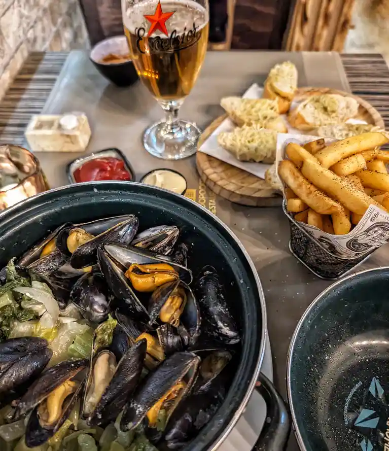 A close-up photo of a table with a large pot of steamed mussels, a basket of french fries, garlic bread, and a glass of…