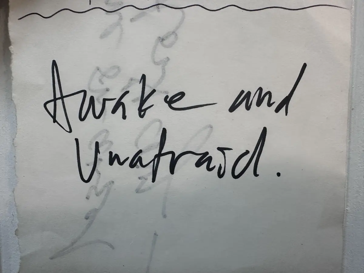 A close-up of handwritten text in black ink on textured white paper, reading 'Awake and Unafraid.' with faint pencil…