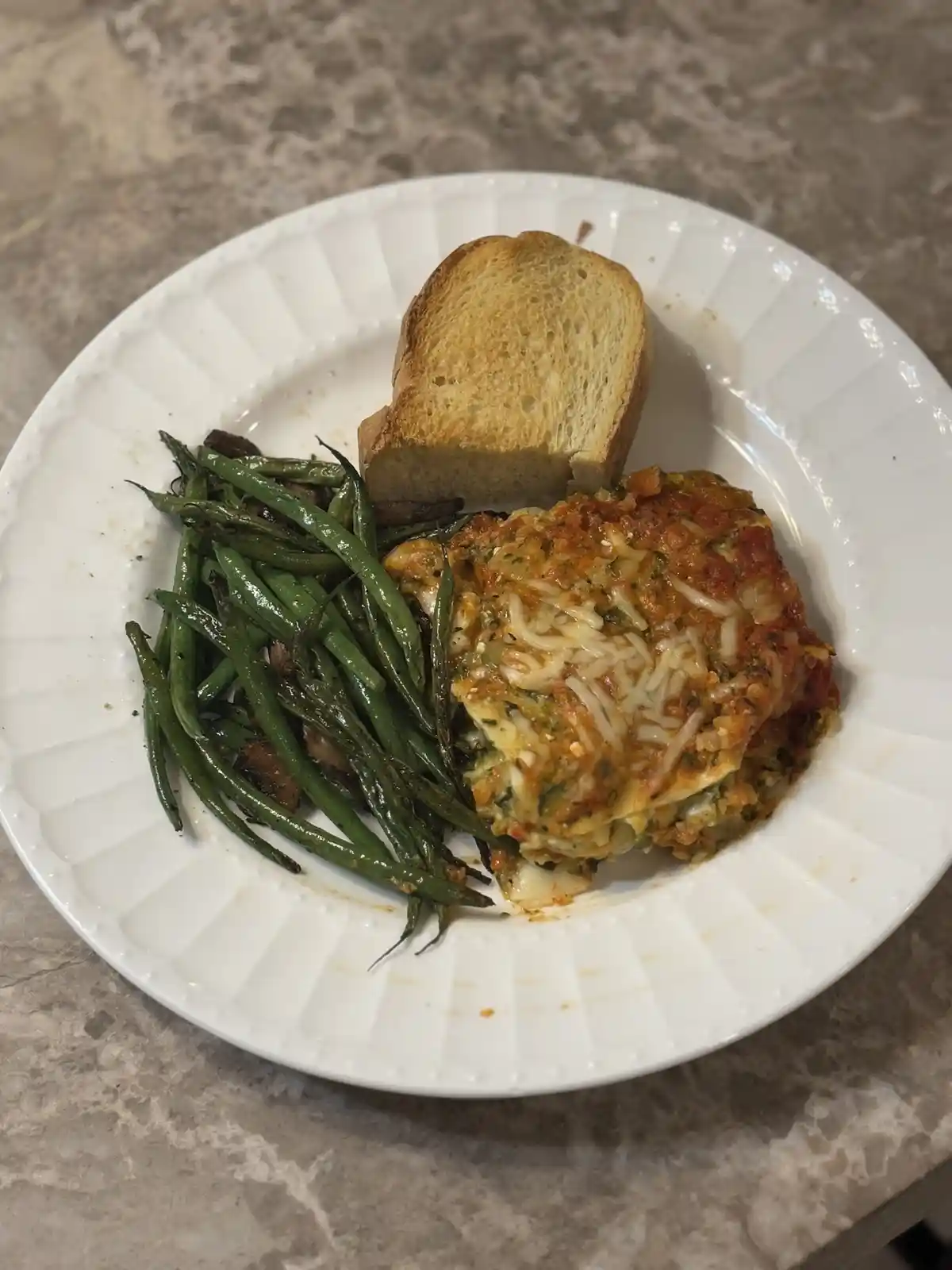 A white plate holds a serving of lasagna, roasted green beans, and a slice of toasted bread on a marble countertop. The…