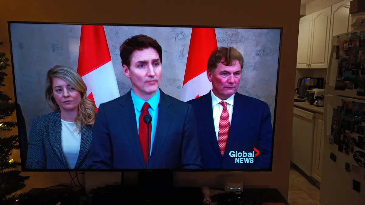 A television screen displays a news broadcast featuring three formally dressed individuals standing before Canadian flags…