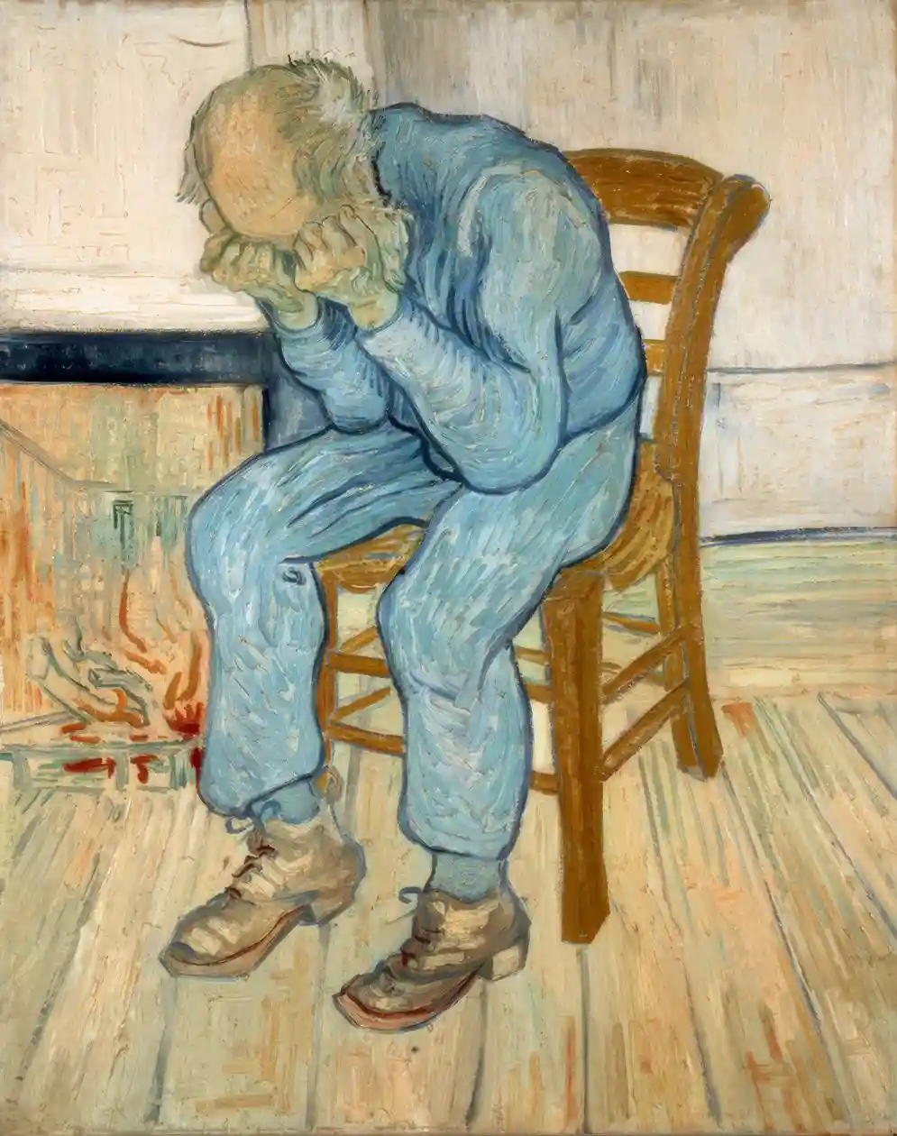 A man in a blue outfit sits on a wooden chair with his head in his hands, appearing distressed. The background shows a…