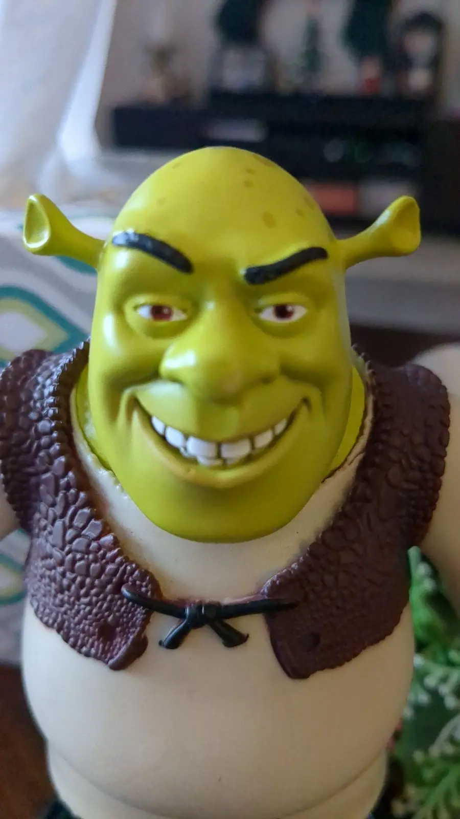 A close-up photo of a green ogre action figure, resembling Shrek, with a wide, toothy grin and furrowed brows, wearing a…