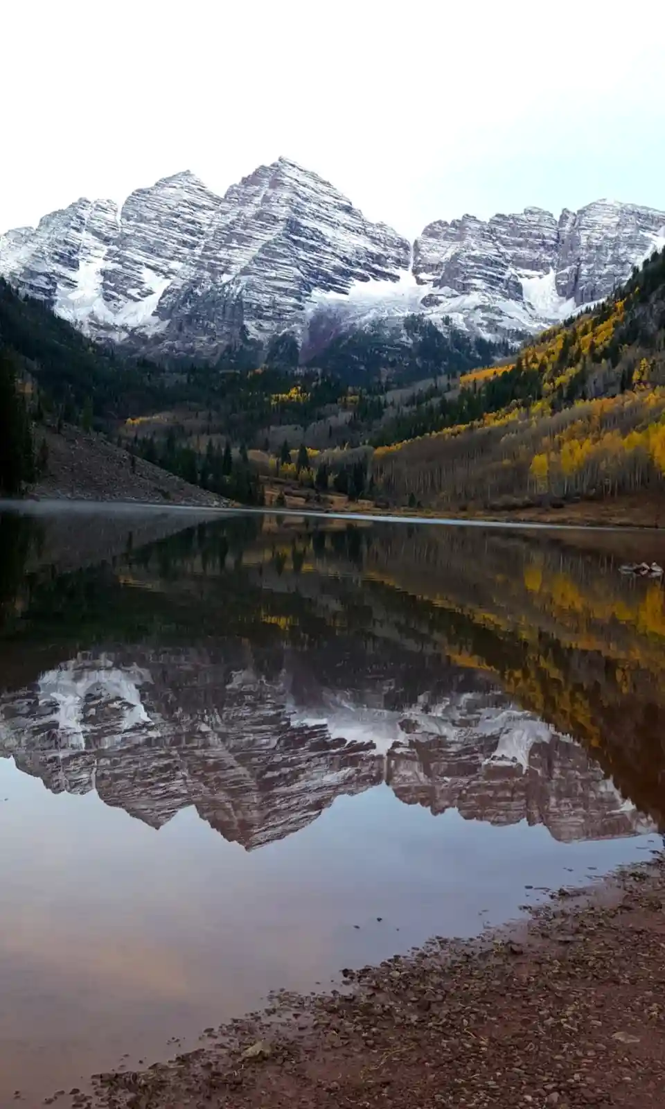A serene landscape photo shows snow-capped mountains reflected perfectly in a calm lake, surrounded by autumn-colored trees…
