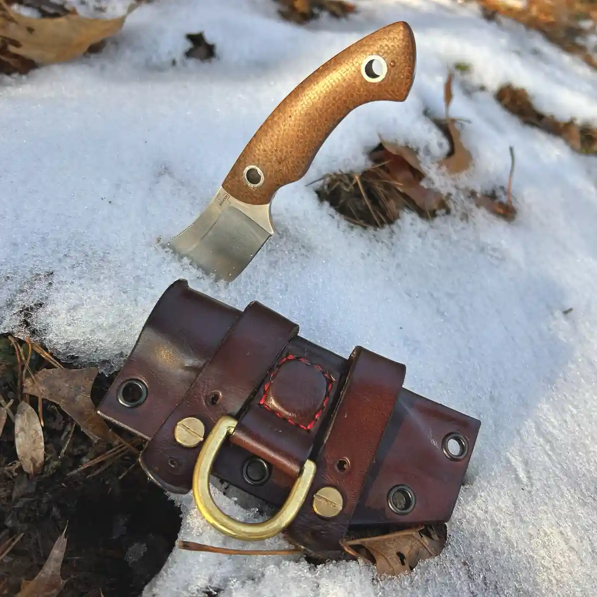 A close-up photo shows a fixed-blade knife with a brown textured handle and silver blade stuck upright in a patch of snow…