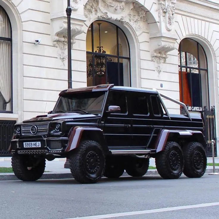a black, heavily modified six-wheeled Mercedes-Benz G-Class SUV with a custom body kit and large off-road tires is parked on…