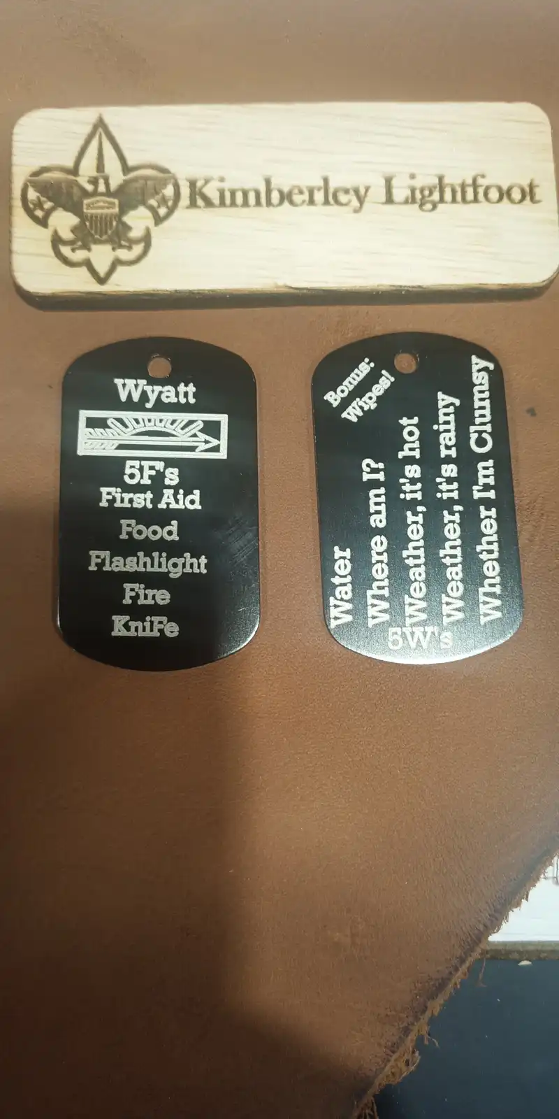 Two black dog tags sit below a wooden plaque engraved with a Scout emblem and the name 'Kimberley Lightfoot'. The left tag…