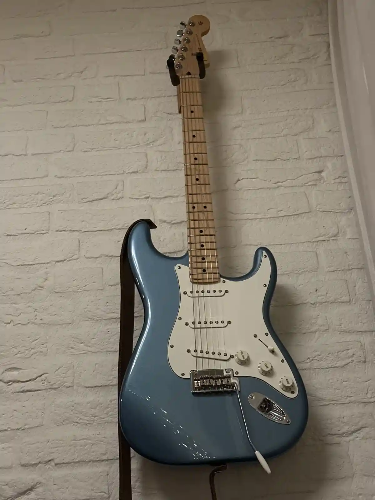 A light blue electric guitar with a white pickguard and natural wood fretboard is mounted vertically against a white brick…