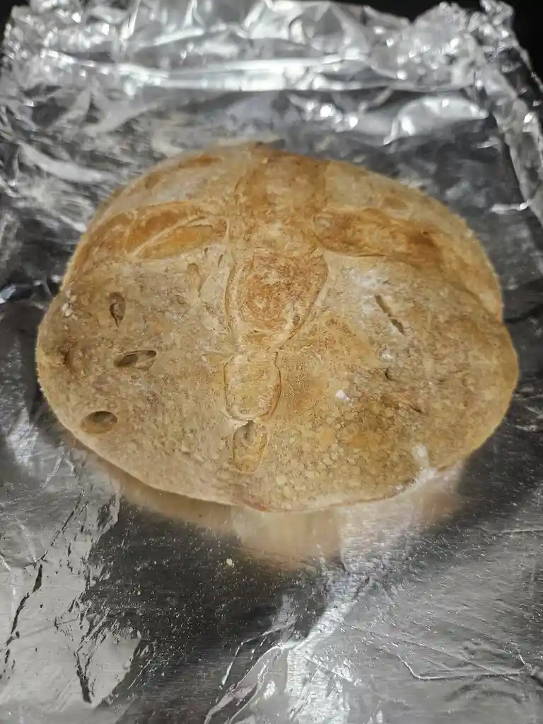 A round, golden-brown loaf of bread with a cracked, textured surface sits on crumpled aluminum foil. The bread's pattern of…