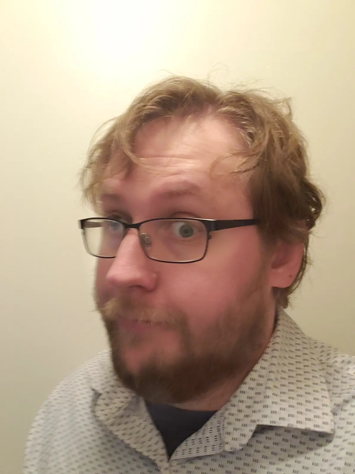A man with glasses and a beard stares with wide, startled eyes at something off-camera. He wears a patterned collared shirt…