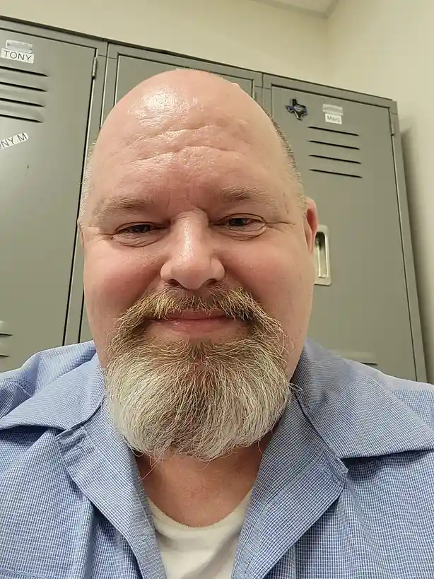 A close-up selfie of a bald man with a full, graying beard and mustache, wearing a blue checkered shirt over a white…