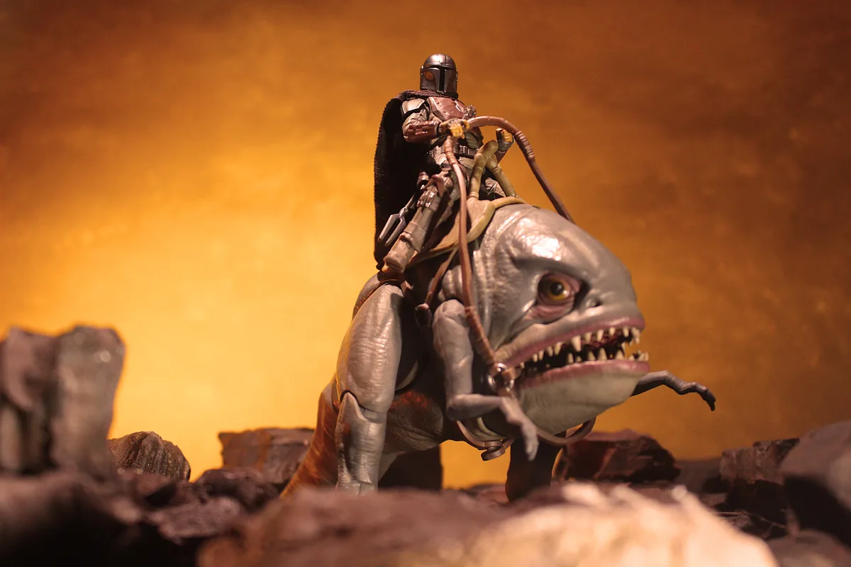 A toy figure of The Mandalorian, wearing his signature helmet and armor, is mounted on a toy of a large, fish-like creature…