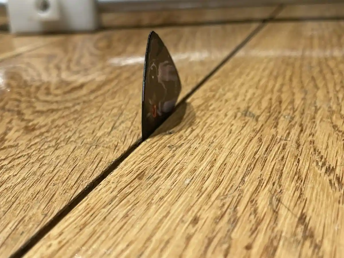 A dark, triangular guitar pick with a printed design is standing upright in the seam between two wooden floorboards. The…
