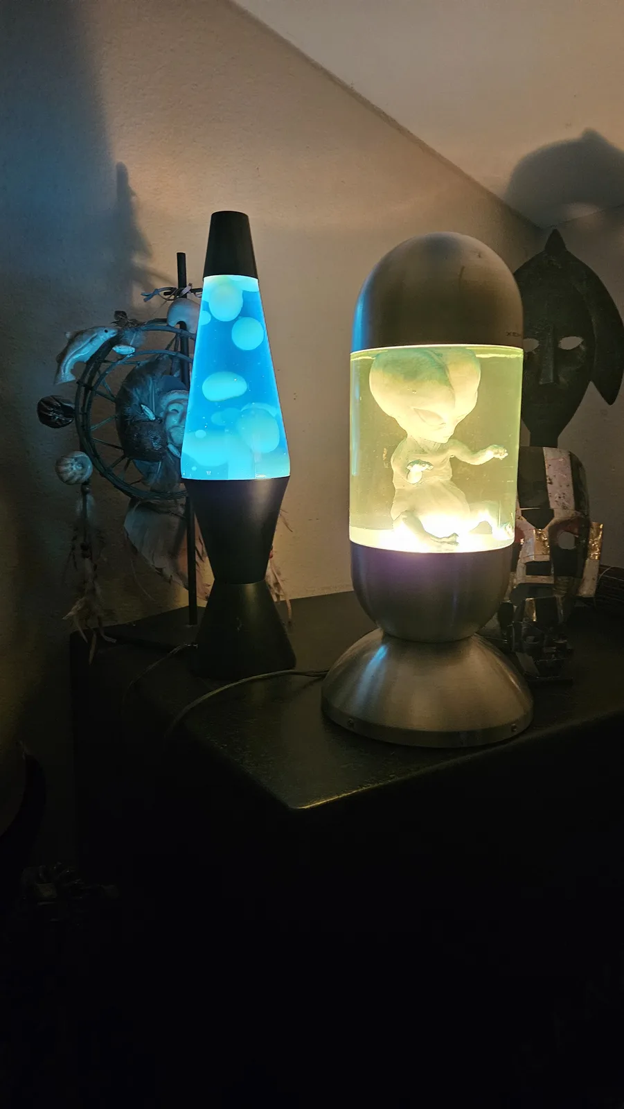 Two lava lamps sit on a dark surface; the left is blue with floating orbs, the right contains a glowing alien baby figurine…
