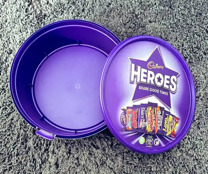 An open purple Cadbury Heroes tin sits on a grey carpet, revealing an empty interior while the lid displays the brand logo…