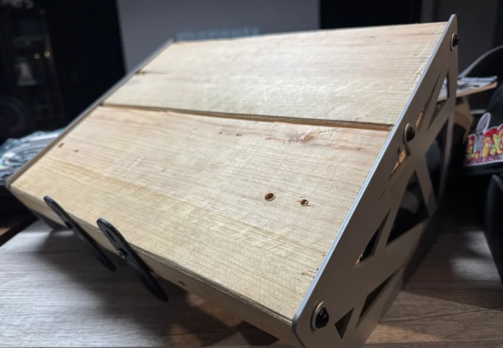 A close-up photo of a homemade wooden monitor stand with a light-colored wood top and a metal frame with cutouts, resting on…