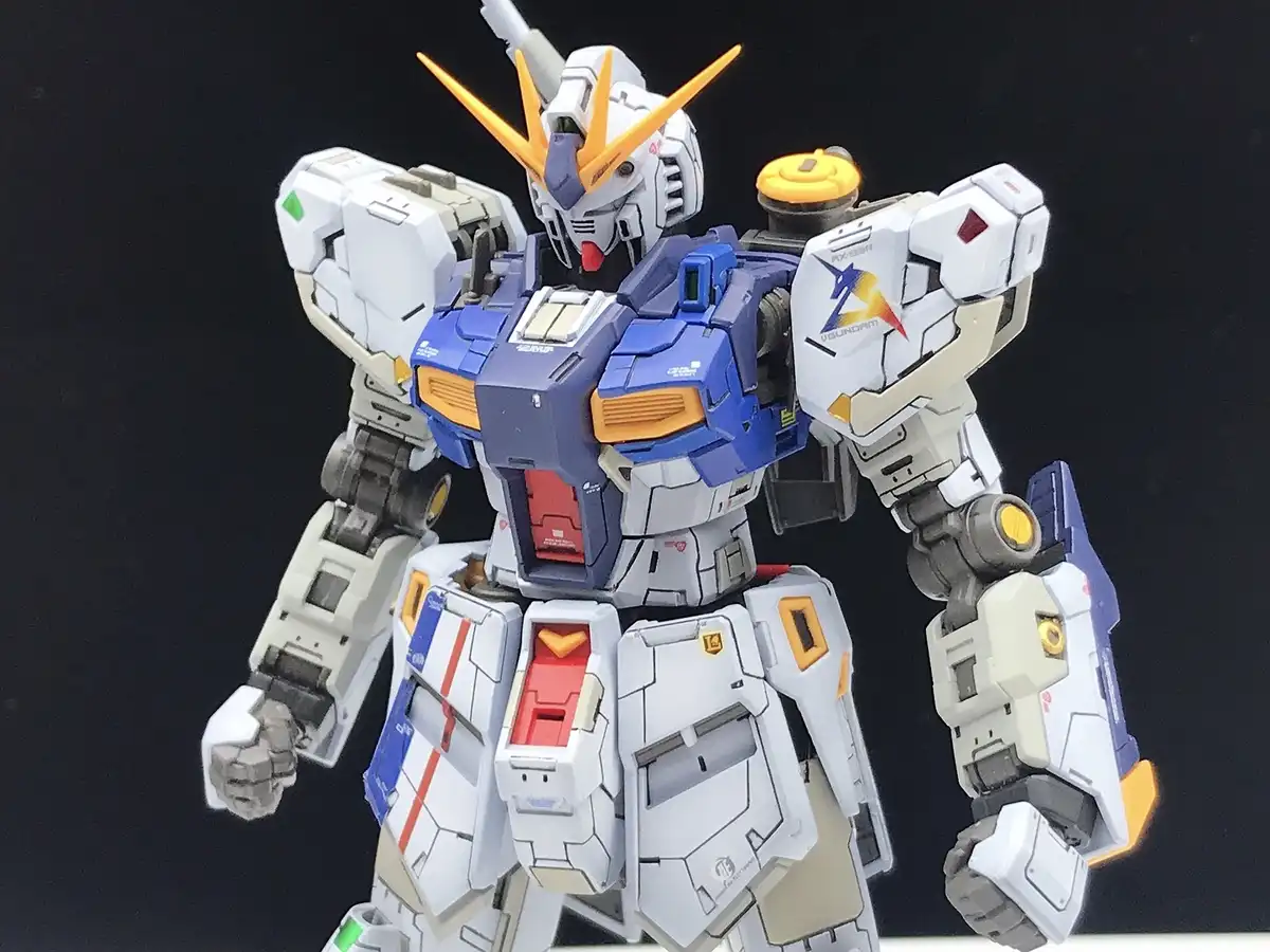 A detailed white, blue, and orange Gundam model stands against a black background with its fists clenched. The text overlay…