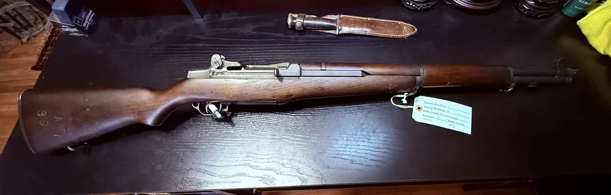 A wooden-stocked rifle lies horizontally on a dark wooden table, with a leather sheathed knife beside it. A white tag…