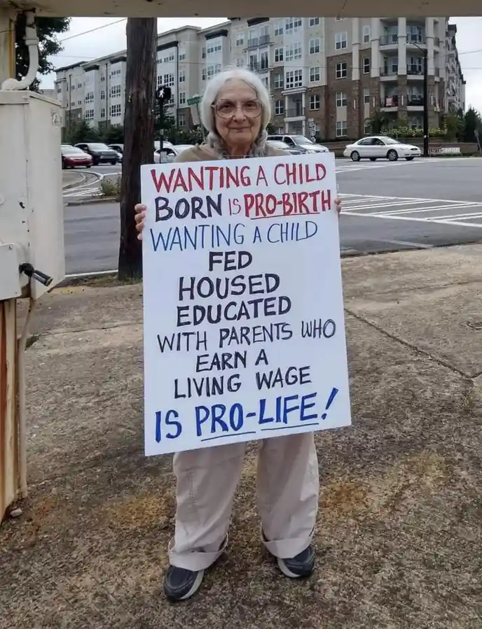 An elderly woman with white hair and glasses stands outdoors holding a handmade white sign with red and blue handwritten…