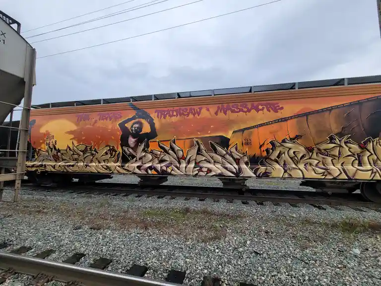 A side view of a freight train car covered in graffiti art depicting a masked figure holding a chainsaw against a fiery…