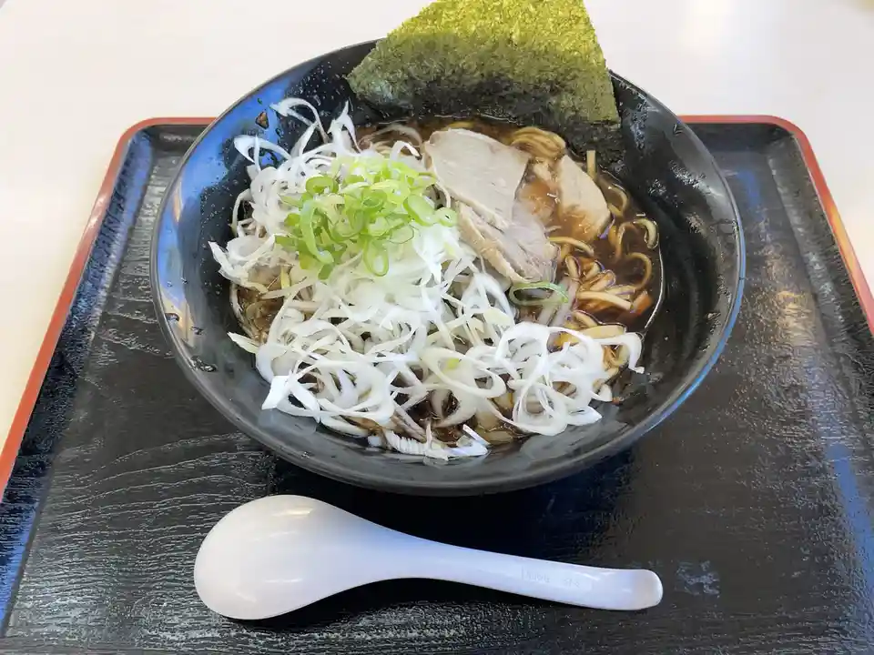 a close-up photo of a black bowl of ramen on a black tray with a white spoon beside it, featuring noodles, broth, sliced…
