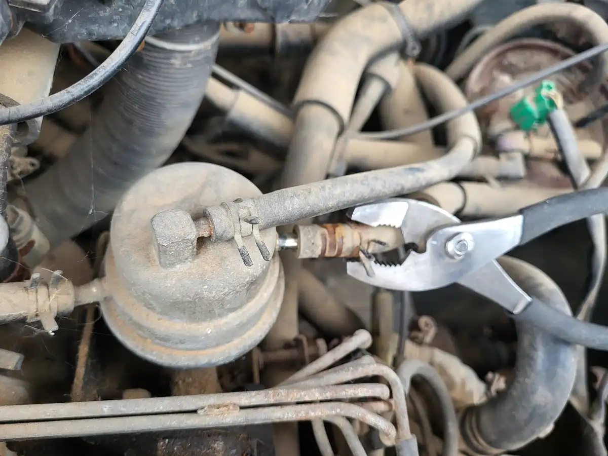 A close-up photo of a dirty, rusty car engine bay with a pair of pliers gripping a corroded metal tube connected to a round…