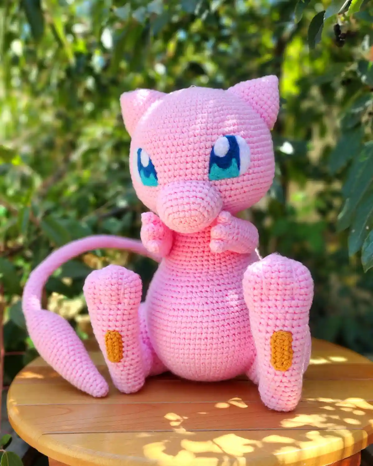 A pink crocheted Mewtwo plush toy sits on a wooden table outdoors with green foliage in the background. The plush has large…