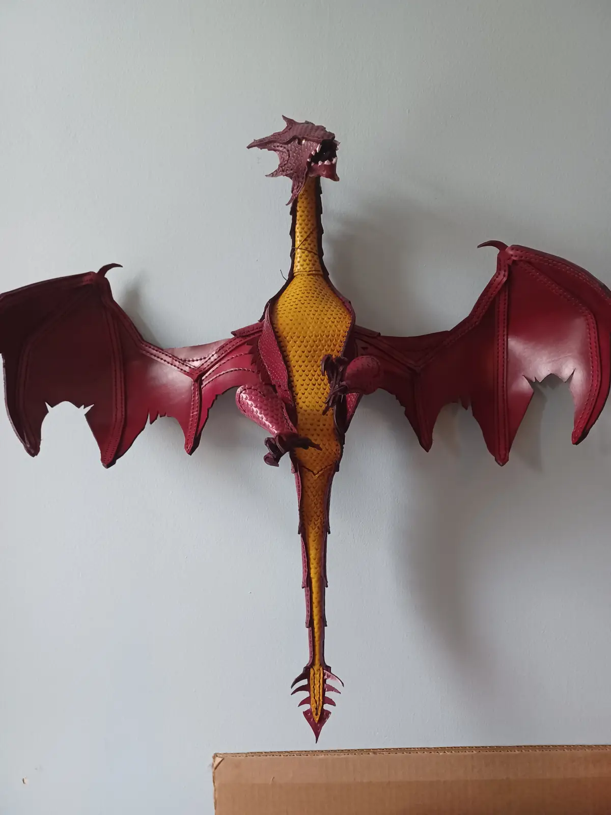 A handmade dragon sculpture mounted on a wall, featuring a yellow LEGO body with red leather wings, head, and tail, with its…